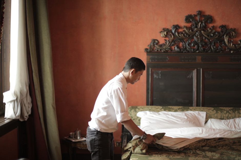 Side view of young male housekeeper making bed while arranging stylish hotel bedroom with vintage interior