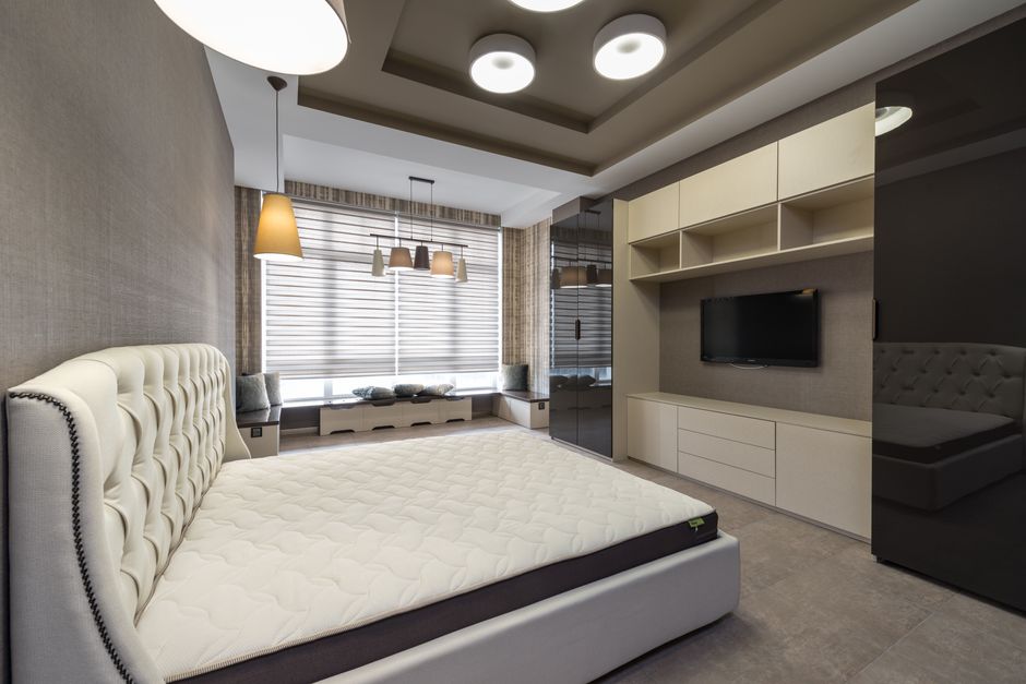 Bed with white mattress placed against wall with cupboards and modern TV in spacious bedroom with window and glowing lamps